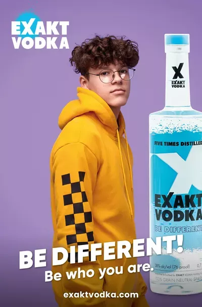 Ein junger Mann in einem gelben Kapuzenpullover vor violettem Hintergrund, der neben einer Flasche Wodka steht.